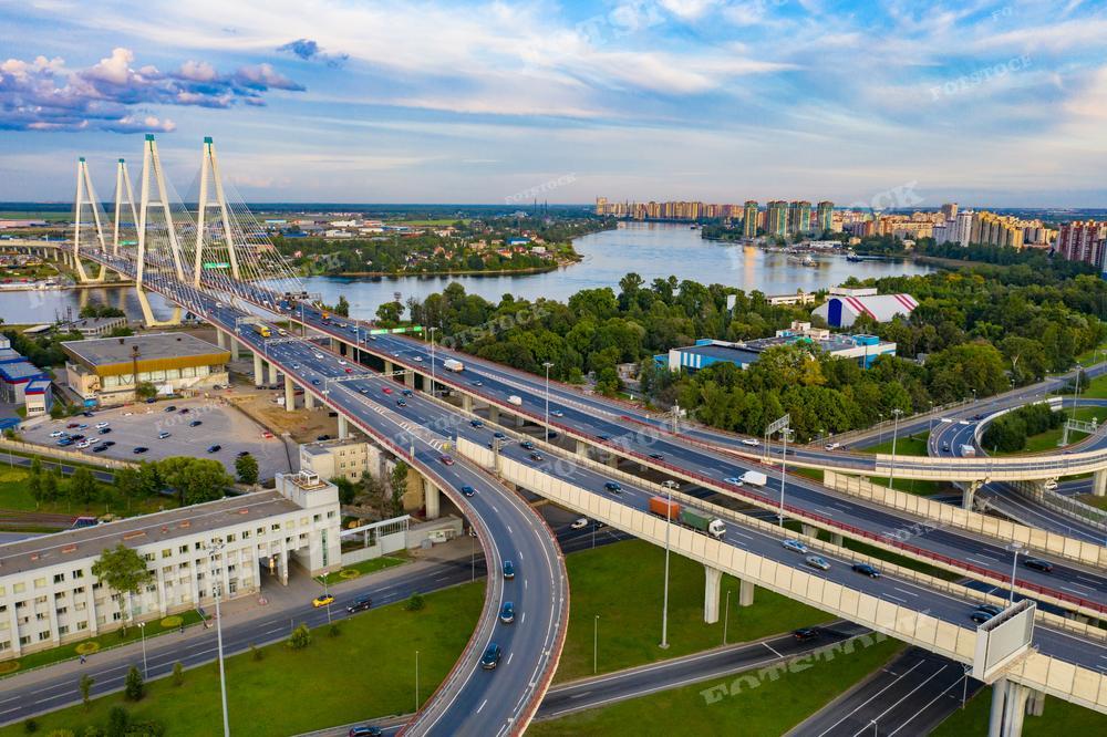 На фотографии изображён современный вантовый мост через широкую реку в городской местности. Мост объединяет два берега, а вокруг него раскинулся живописный пейзаж с жилыми районами, зелёными насаждениями и просторным водоёмом. Фото может быть использовано в градостроительных проектах, для демонстрации примеров современной инфраструктуры, в туристических путеводителях и как элемент дизайна в печатных и электронных изданиях.
